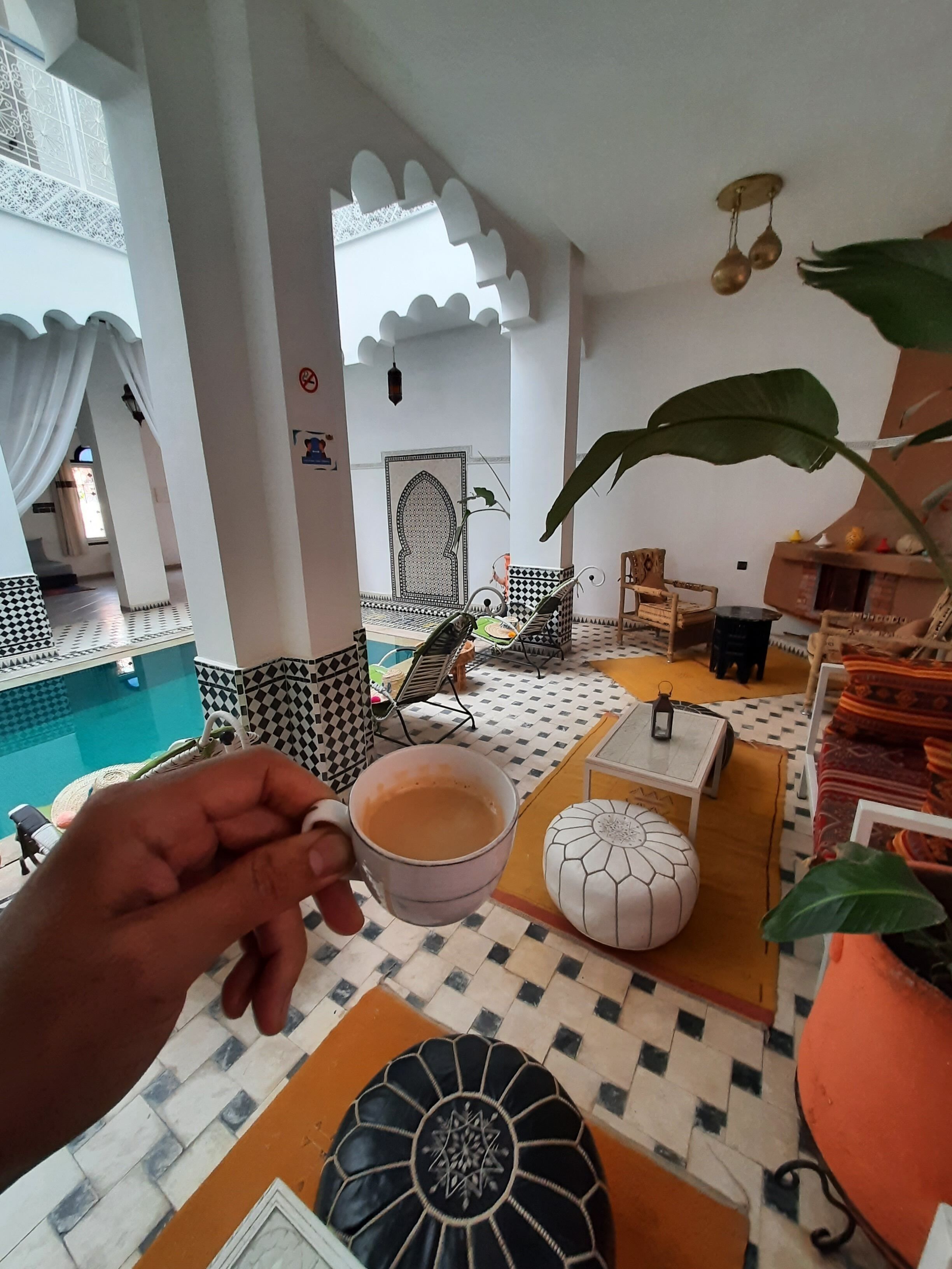 Eine Hand hält eine Tasse, im Hintergrund ein Pool und ein Zimmer mit gemustertem Boden und Möbeln.