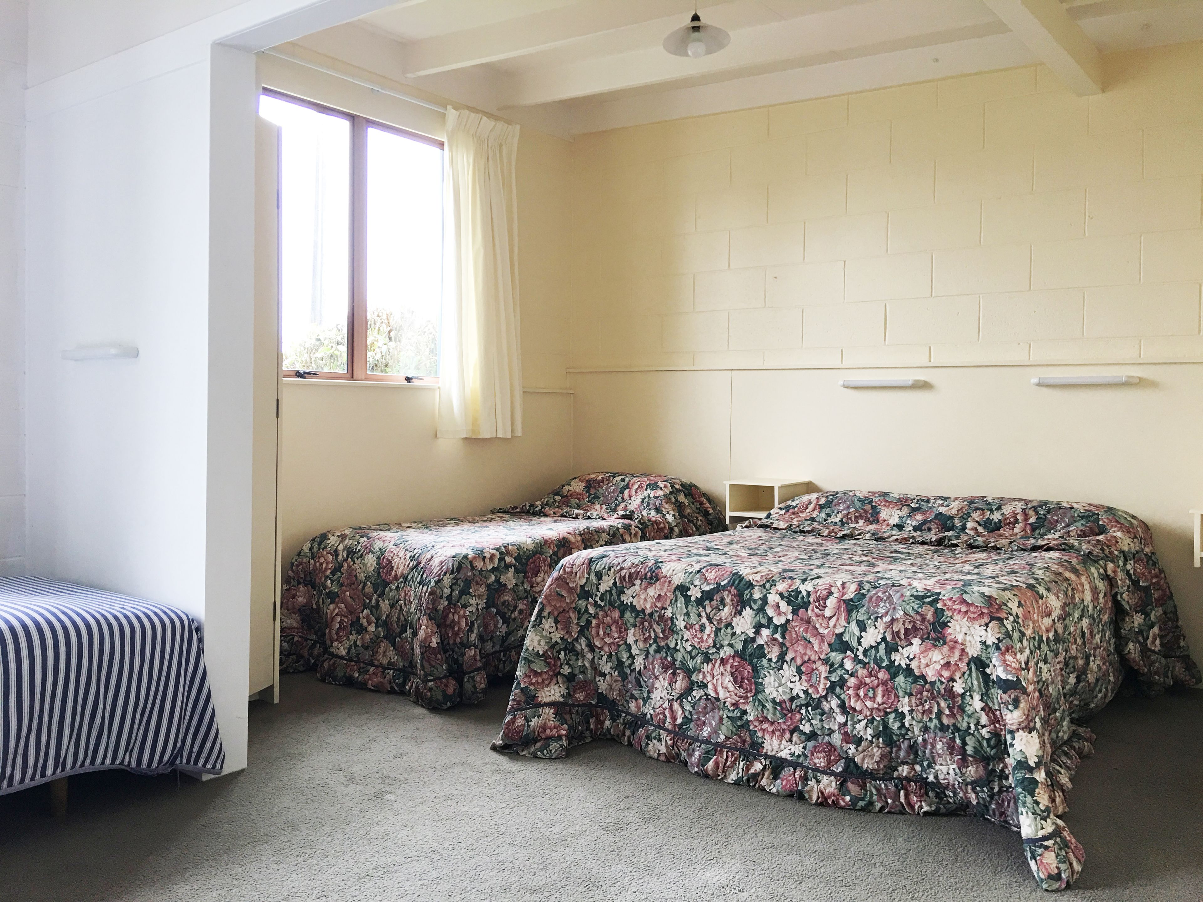 A room with two beds, a bench, a window with curtains, and a brick-patterned wall.