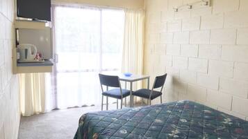 Una habitación pequeña y sencilla con una cama, una mesa con dos sillas, un televisor en un estante y una ventana con cortinas.