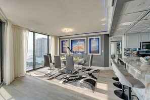Uma zona de refeições moderna com uma mesa grande e cadeiras, uma cozinha com bancada de mármore e vista para o horizonte urbano através de grandes janelas.