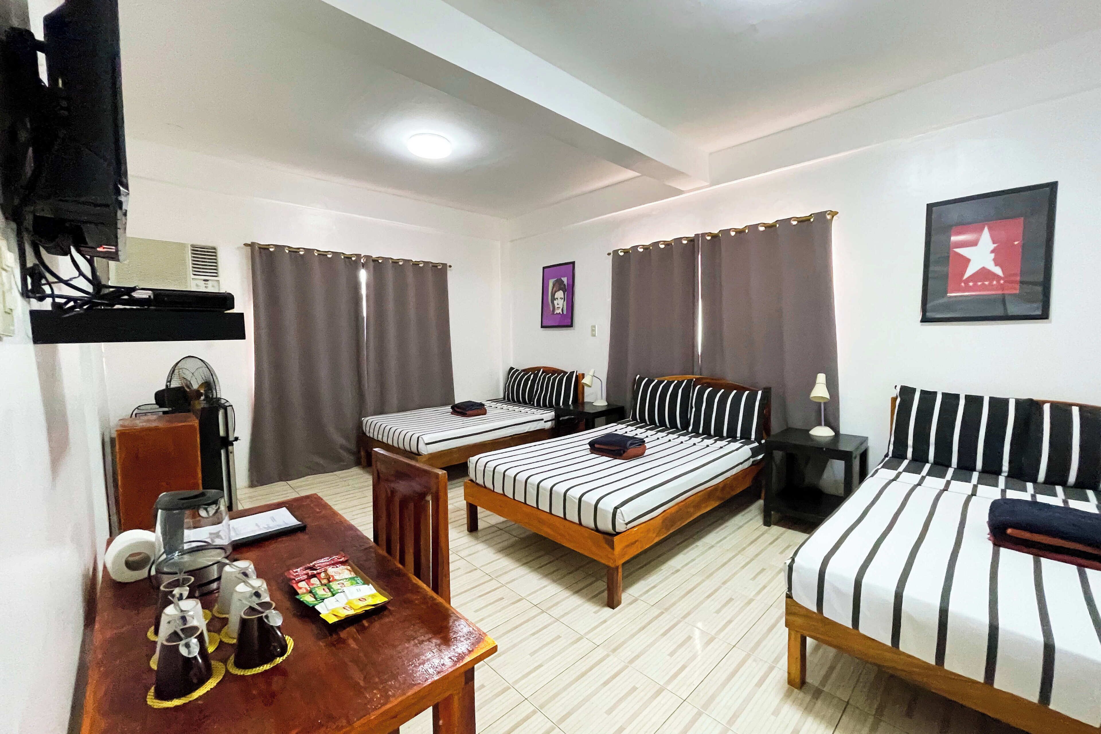 A hotel room with two single beds, a desk with a chair, a TV, a fan, and a small table with cups and a box.
