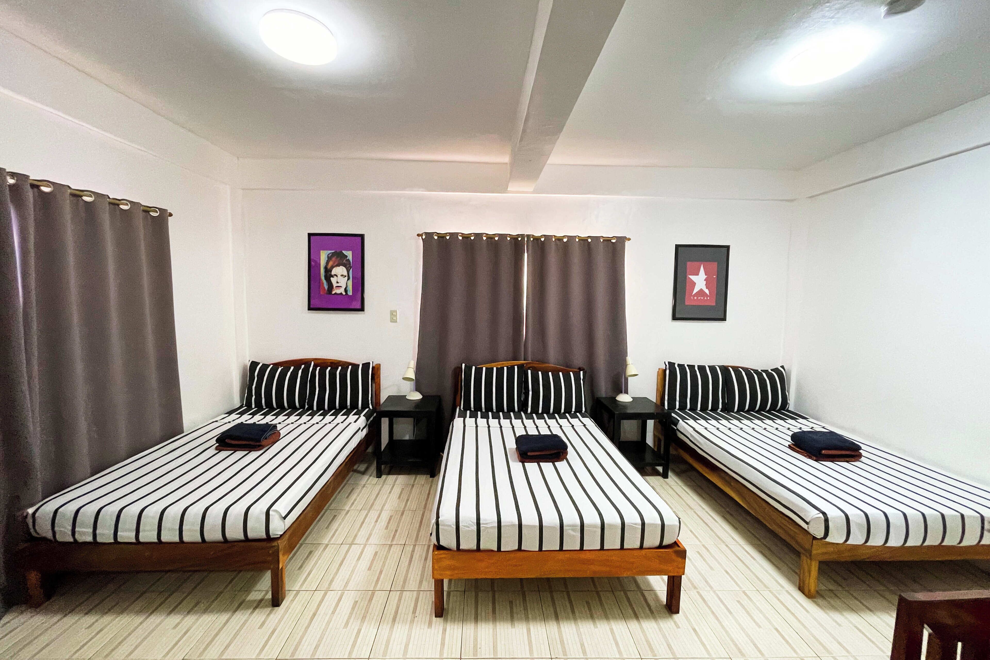 Ein Zimmer mit drei Einzelbetten, jedes mit einem Holzrahmen und gestreifter Bettwäsche. Zwei kleine Tische mit Lampen stehen bereit, und die Wände sind schlicht gehalten, mit zwei gerahmten Bildern an den Wänden.