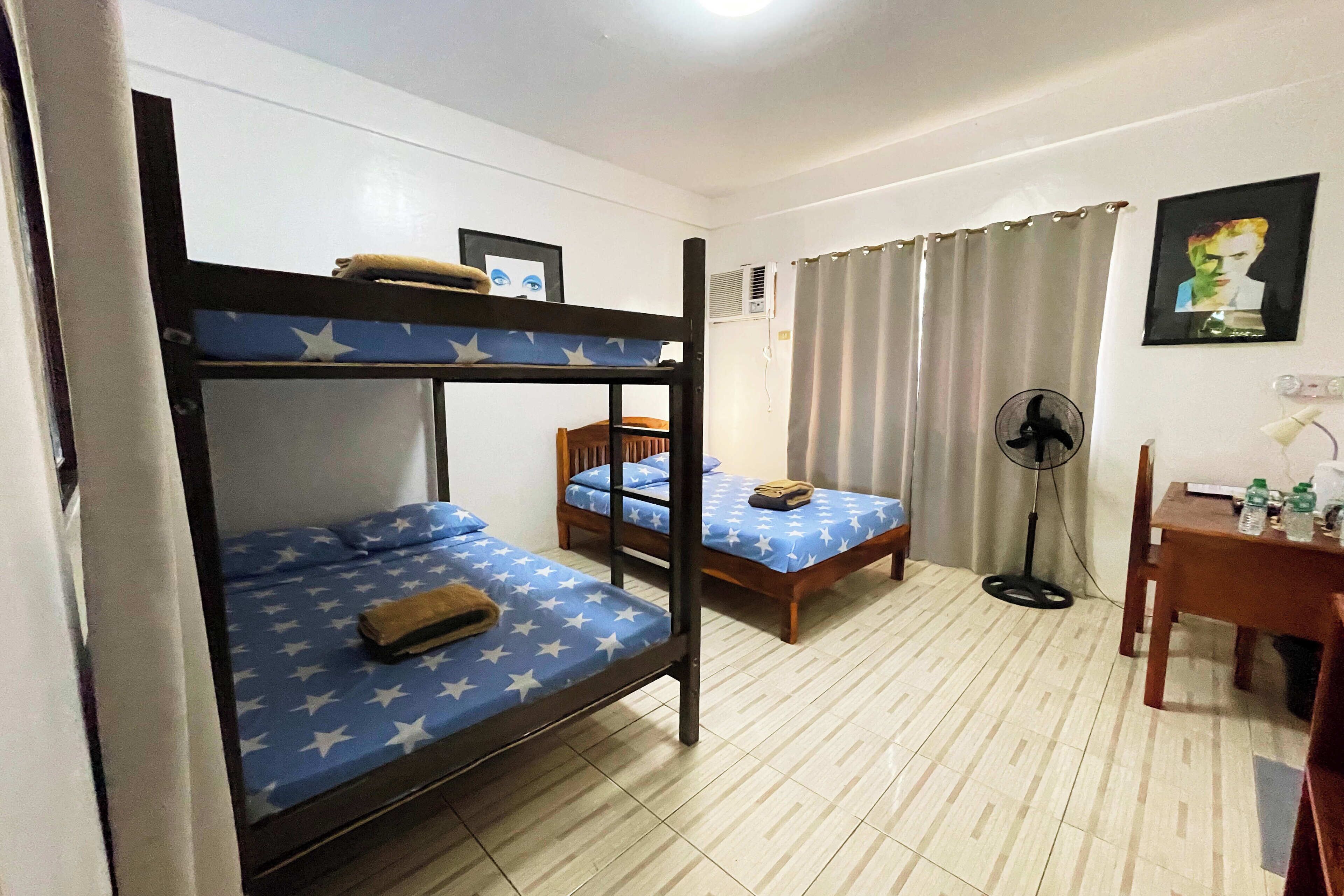 A room with bunk beds, a desk, a chair, a fan, and a picture on the wall.