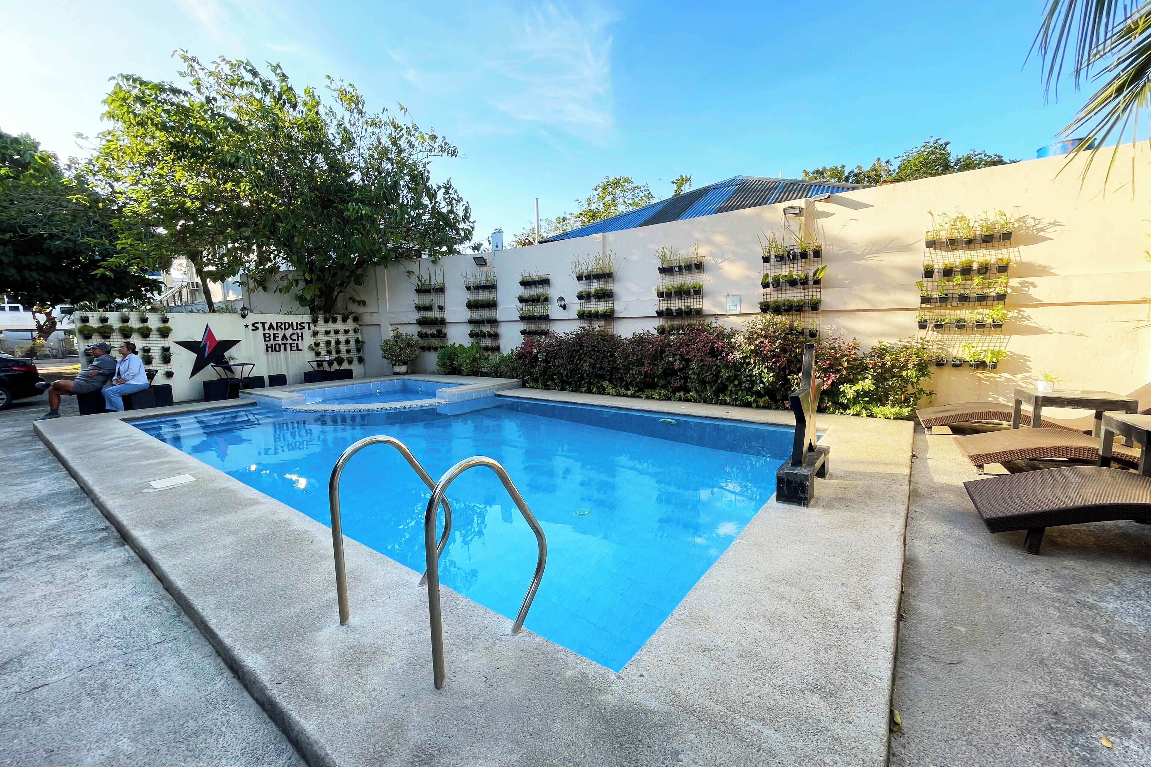 Una piscina con una escalera metálica, rodeada de sillas de descanso y un muro con jardineras colgantes.