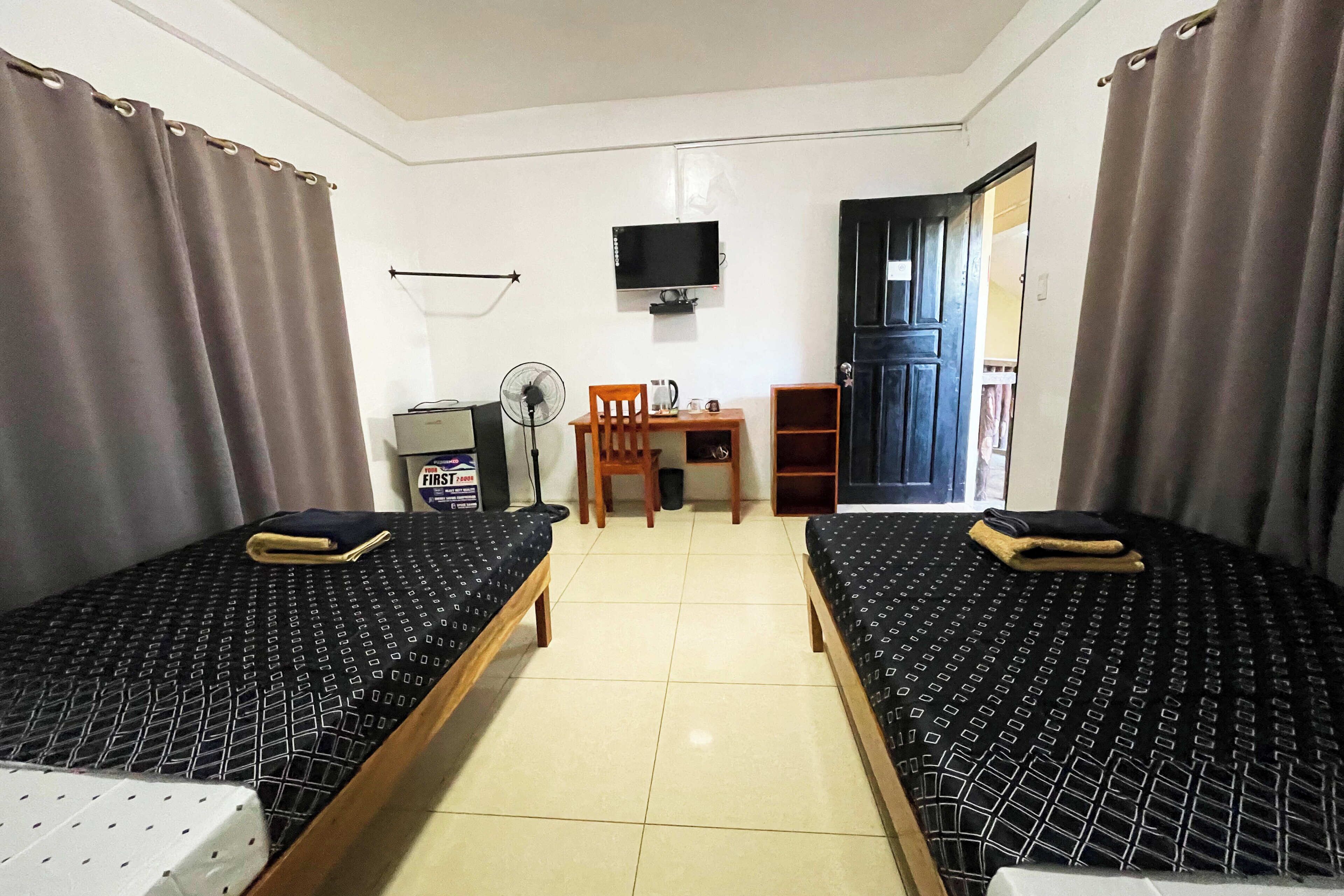 Quarto com duas camas, uma escrivaninha, uma cadeira, uma televisão e uma porta.