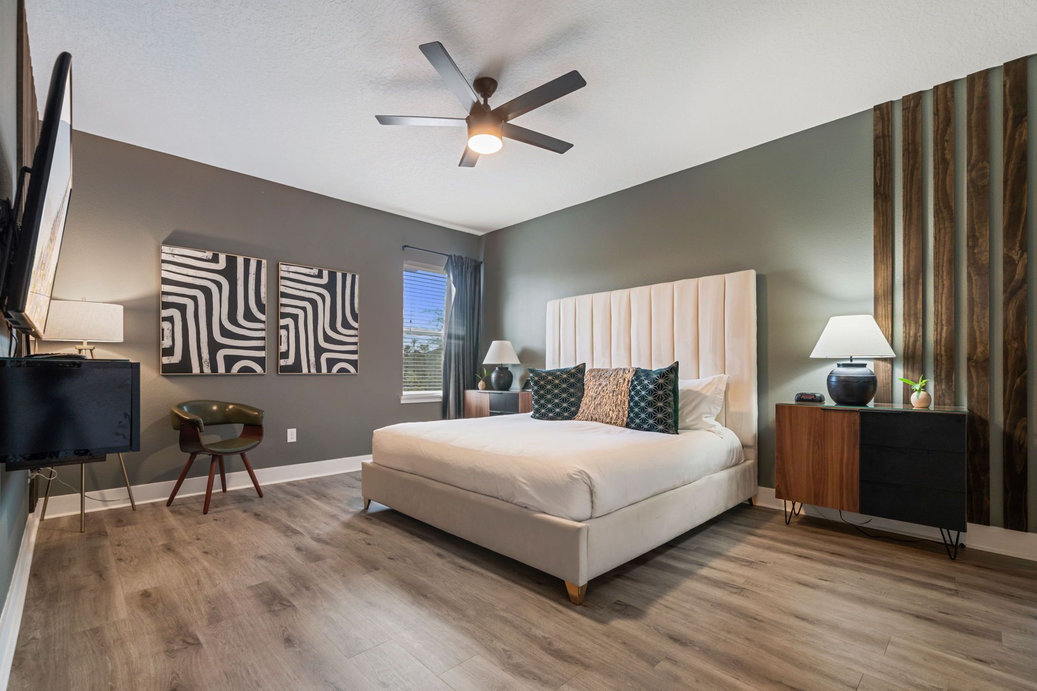 A bedroom with a large bed, a ceiling fan, a TV, and a desk with a lamp.