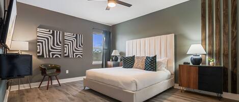 A bedroom with a large bed, a ceiling fan, a TV, and a desk with a lamp.