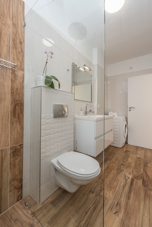Et moderne badeværelse med glasbrusekabine, hvid vask, toilet og indbygget vaskemaskine.