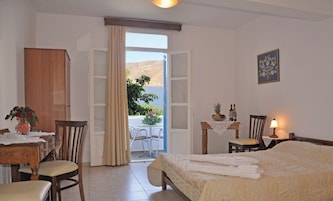 Quarto de hotel com cama, mesa com cadeiras, armário de madeira, varanda com vista para o mar e abajur na mesinha de cabeceira.