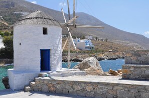 A traditional white building with a blue door, a stone platform, and a windmill in the background. - Filoxenia (Amorgos)
