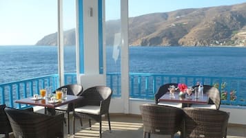 Een terras met rieten meubels en eettafels, met uitzicht op een kustlandschap met heuvels en de zee.
