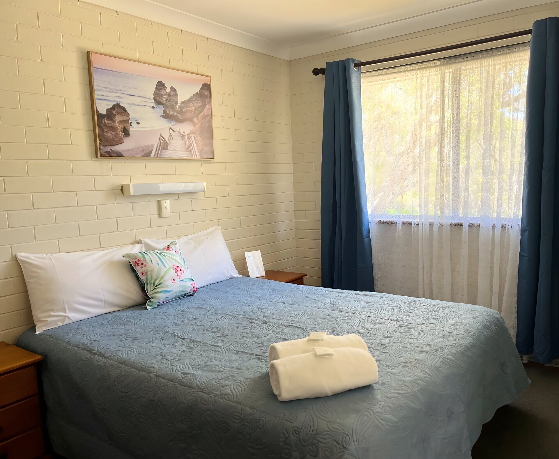 Habitación de hotel con cama, almohadas, un cuadro, ventana con cortinas y mesita de noche.