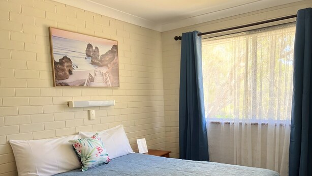 Een hotelkamer met een bed, kussens, een schilderij, een raam met gordijnen en een nachtkastje.