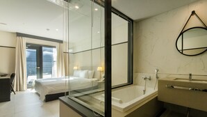 Ein modernes Hotelzimmer mit einem großen Bett, einer in Glas eingefassten Dusche, einem separaten Badezimmer mit Badewanne und Blick auf die Stadt durch das Fenster.