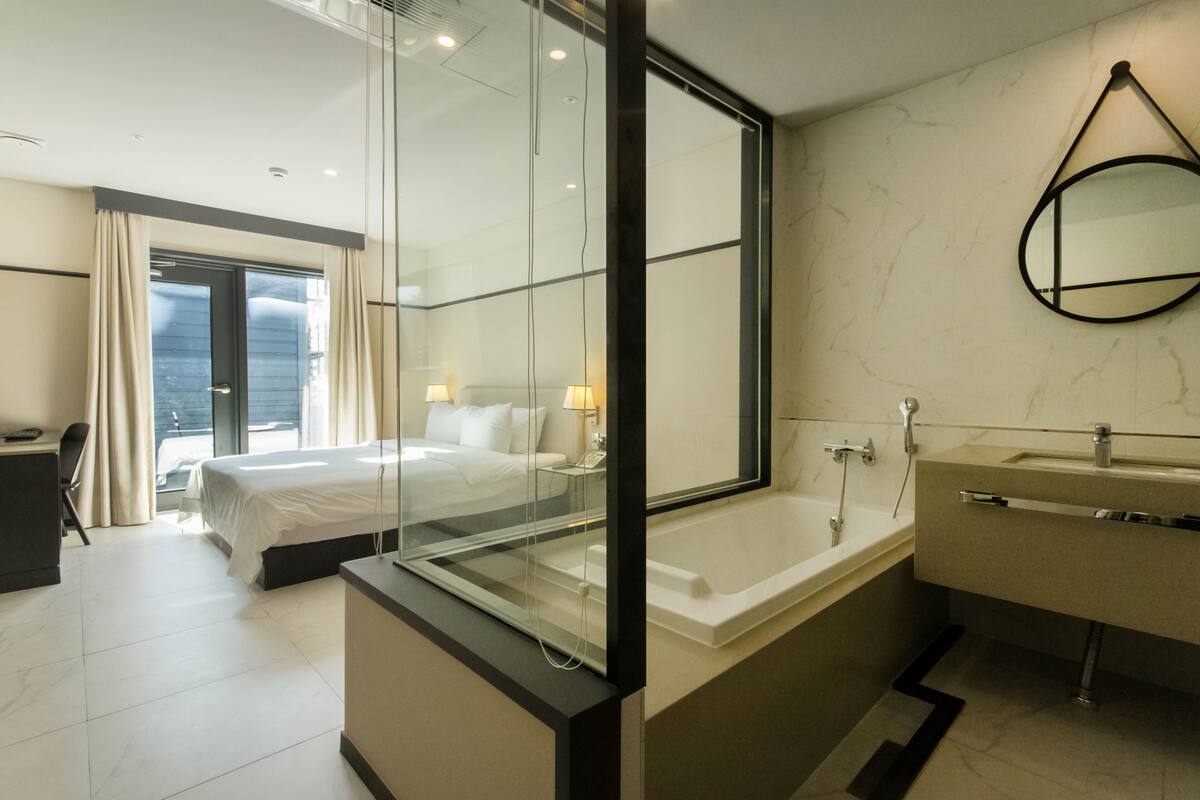 A modern hotel room with a large bed, a glass-enclosed shower, a separate bathroom with a bathtub, and a view of the city through the window.
