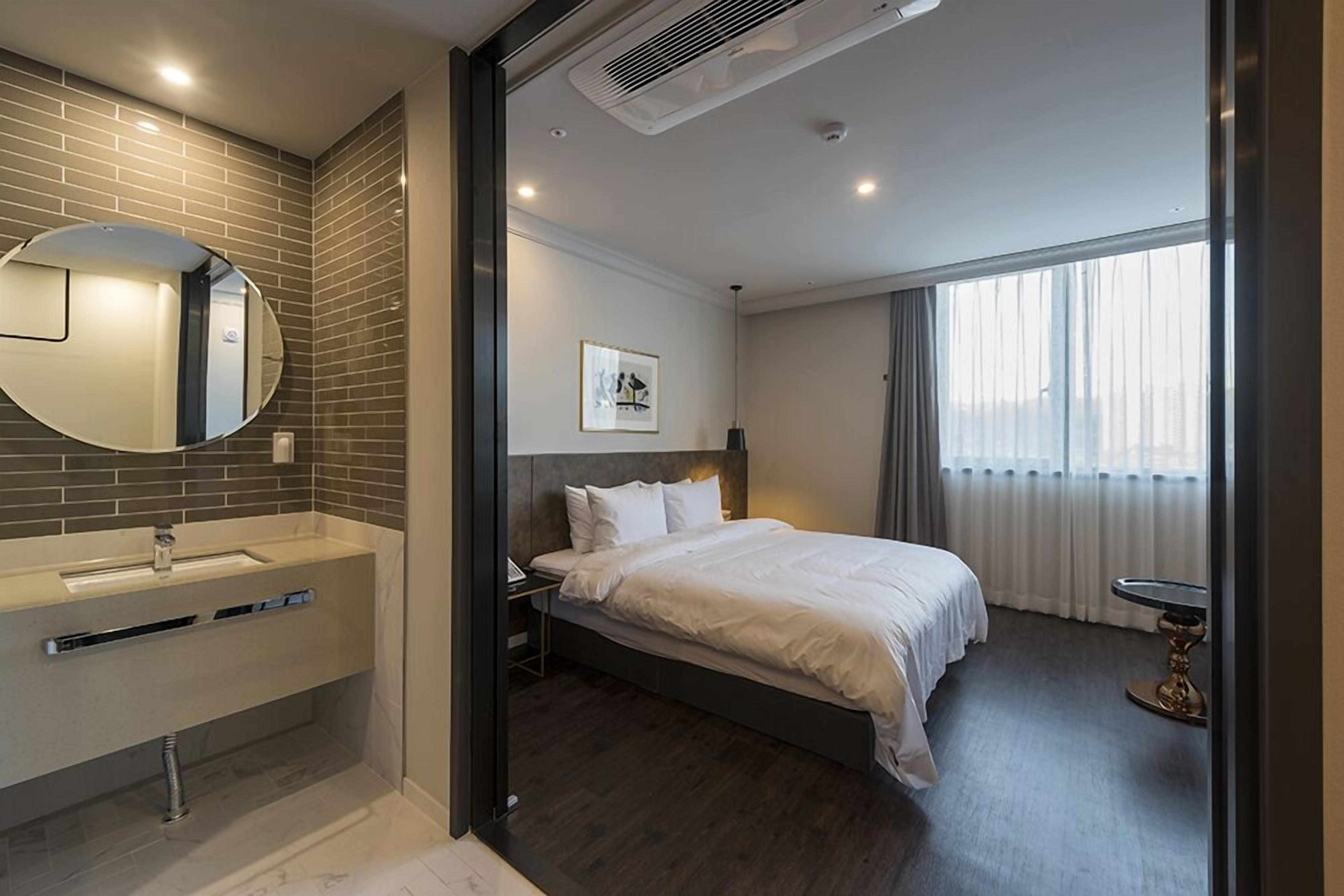 A modern hotel room with a large bed, a round mirror, a built-in sink, and a window with curtains.