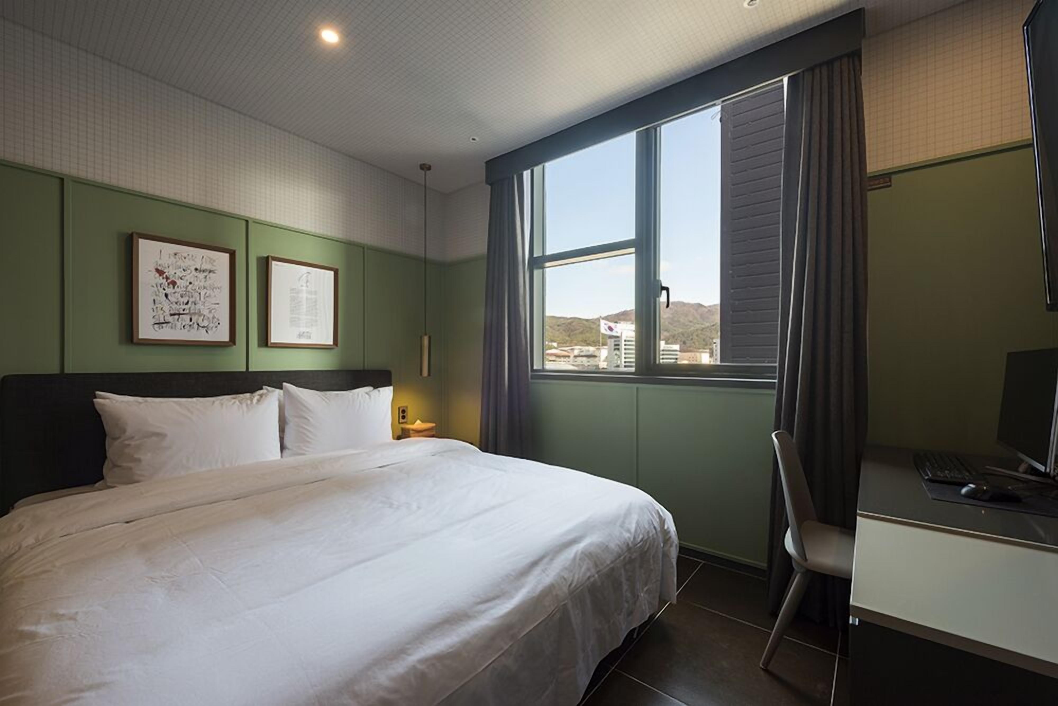 Habitación de hotel con cama, escritorio con computadora, silla y ventana que ofrece vistas a edificios y colinas.