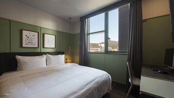 Habitación de hotel con cama, escritorio con computadora, silla y ventana que ofrece vistas a edificios y colinas.