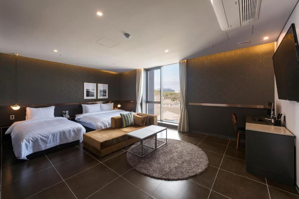 A modern hotel room with two beds, a sofa, a desk, and a large window.