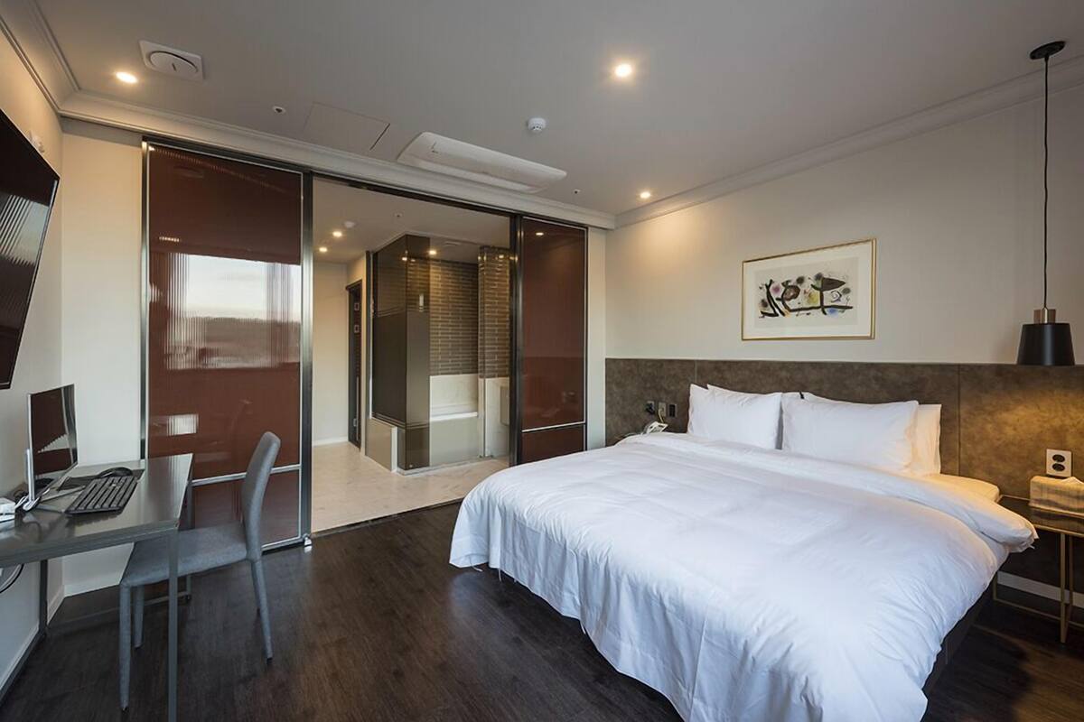 A modern hotel room with a large bed, a desk with a computer, a glass door leading to a bathroom, and a framed artwork on the wall.