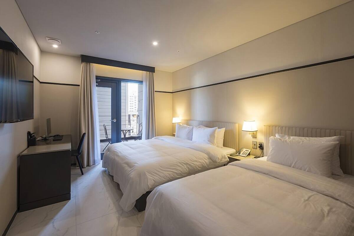 A hotel room with two beds, a desk, a chair, a lamp, and a window with curtains.