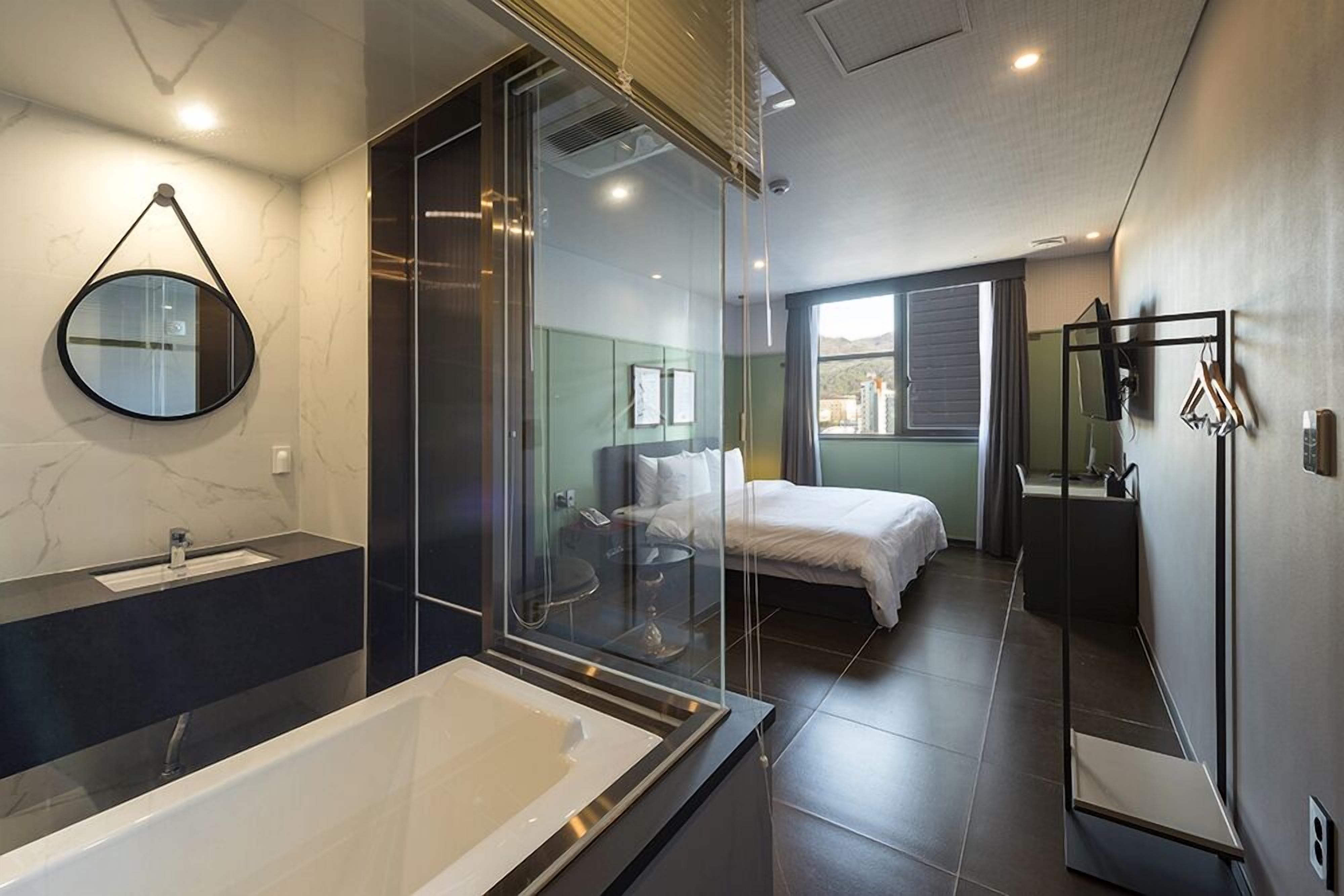 A modern bathroom with a large bathtub, a glass shower enclosure, and a bed visible through the shower.
