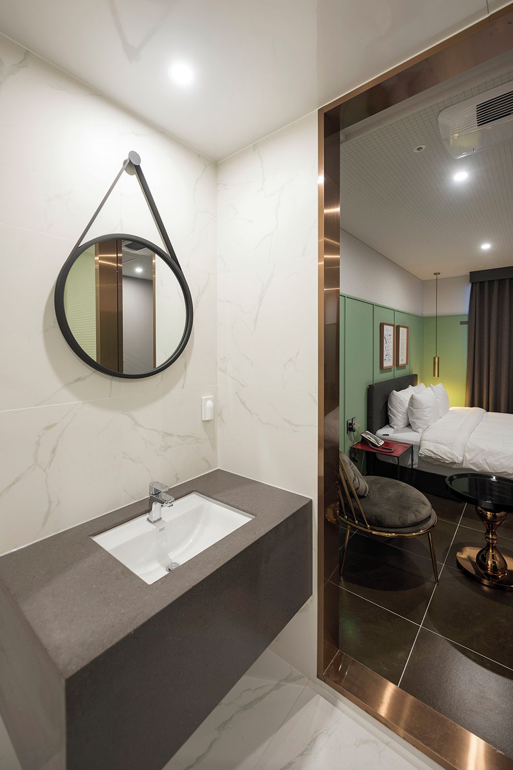 Un baño moderno con un lavamanos de mármol, un espejo redondo y vistas a un dormitorio con cama y sillón.