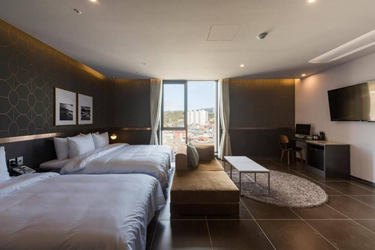 A modern hotel room with two beds, a desk, a TV, and a large window.