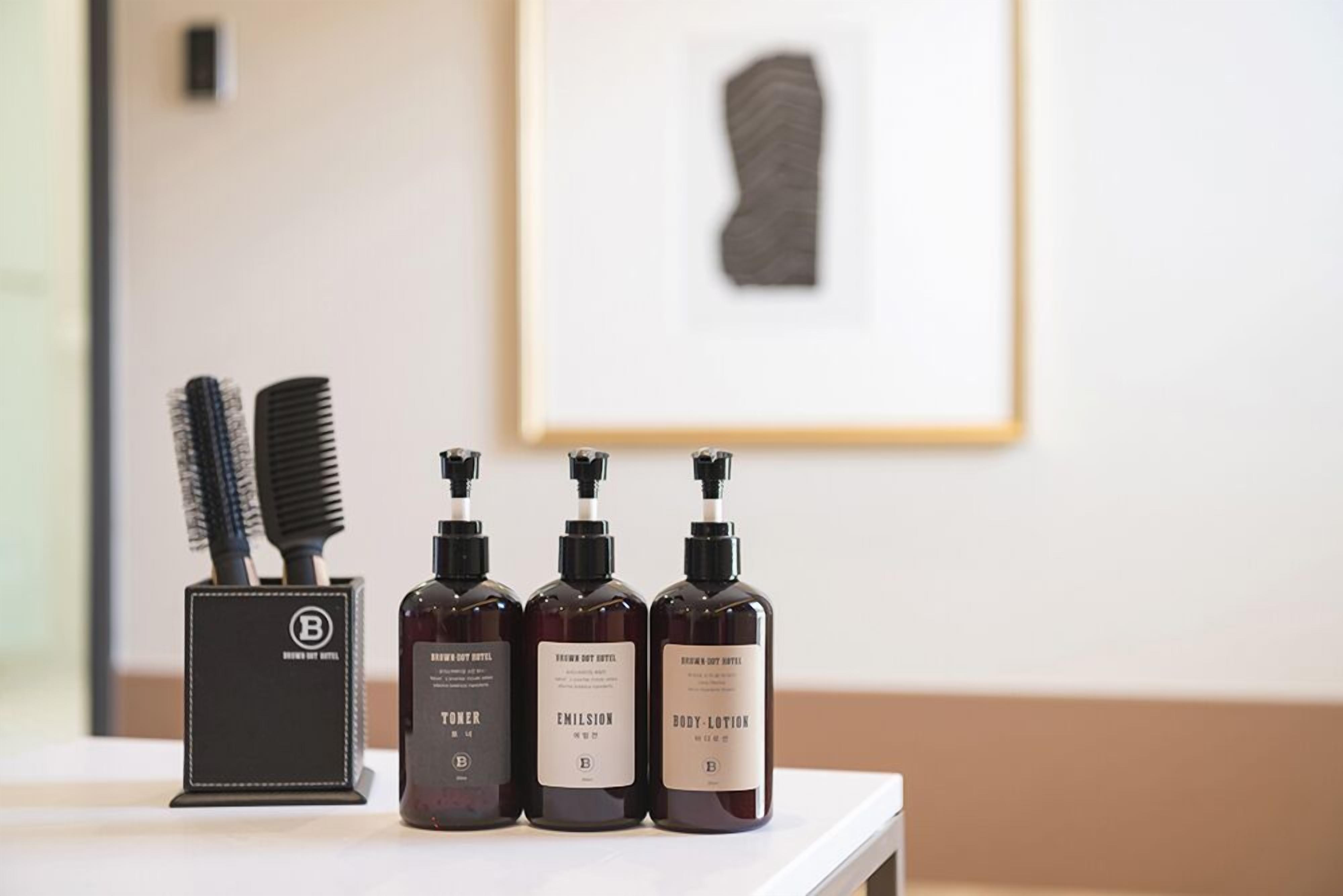 A bathroom counter with a hairbrush holder, three bottles of body lotion, and a framed artwork on the wall.