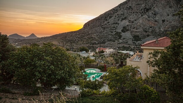 Un paysage montagneux au coucher du soleil, un bâtiment avec un toit recouvert de tuiles rouges et un emblème vert au premier plan.