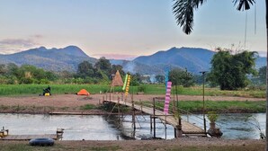 Un camping junto al río, con un puente de madera, tiendas de campaña y una estructura con techo de paja.