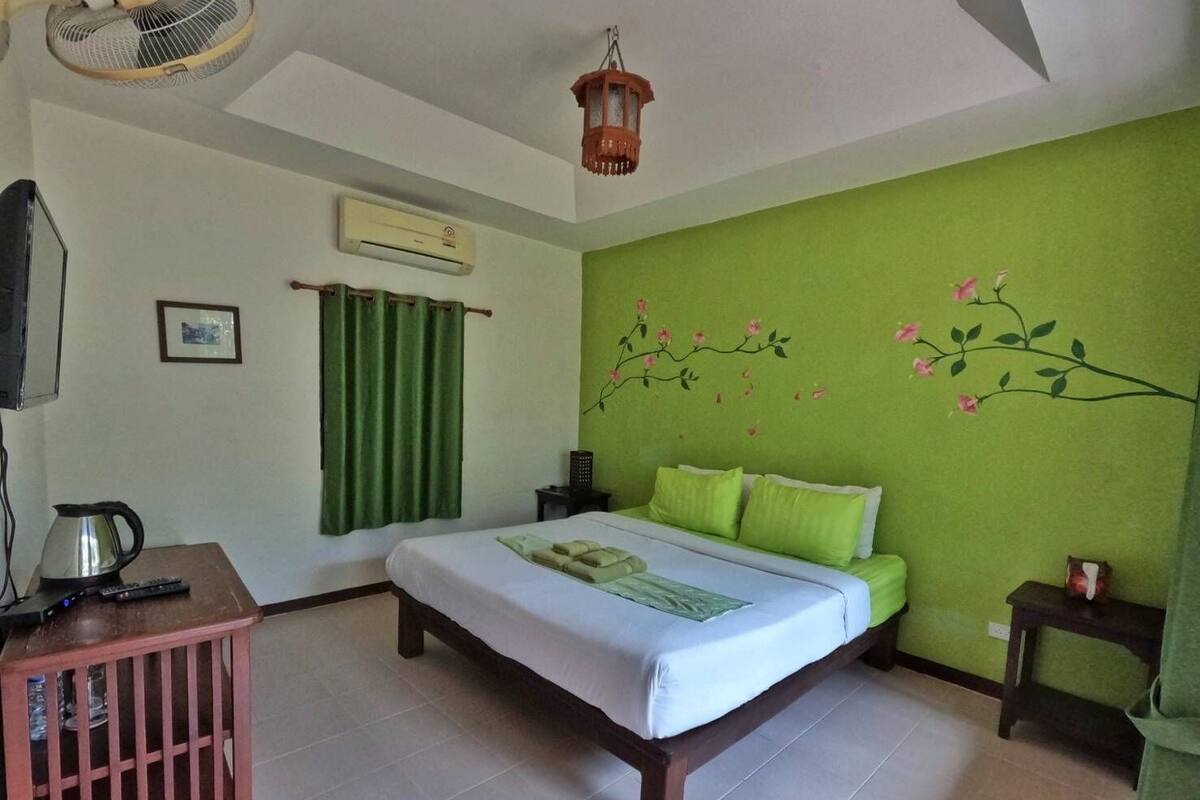 A room with a bed, green curtains, a wall-mounted air conditioner, a small table with a kettle, a fan, and a decorative branch with flowers on the wall.