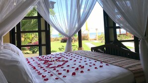 Quarto com cama coberta de pétalas de rosa, vista para um jardim e para o mar através de grandes janelas, e um dossel sobre a cama.