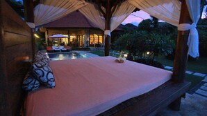 Uma cama com dossel, uma piscina e um edifício com telhado vermelho ao fundo.