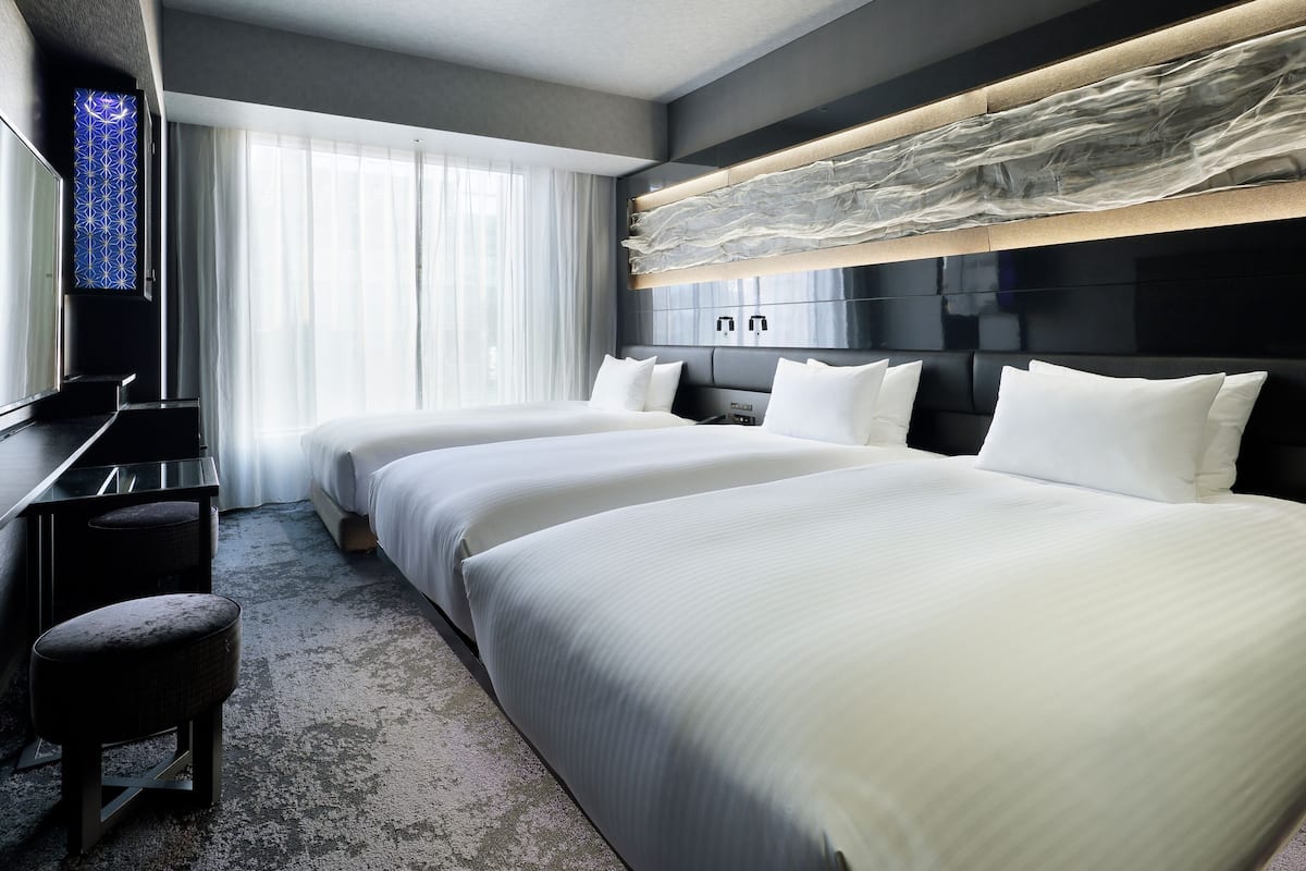 Quarto de hotel com duas camas, uma escrivaninha, uma cadeira e uma janela grande com cortinas leves.