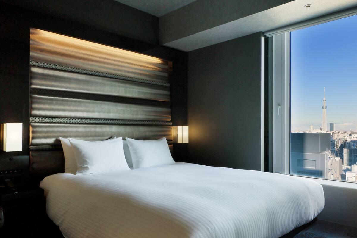 Quarto de hotel com uma cama grande, cabeceira com linhas horizontais, duas luminárias de apoio e vista para a cidade através de uma janela ampla.