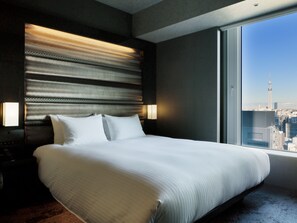 Een hotelkamer met een groot bed, een hoofdeinde met horizontale lijnen, twee bedlampjes en een stadsgezicht door een groot raam.