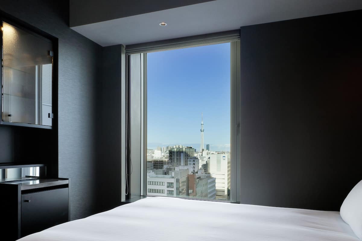 Quarto de hotel moderno com uma cama grande, escrivaninha embutida e vista para a cidade através de uma janela ampla.