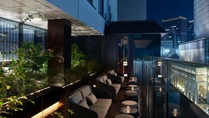 Ein moderner Außen-Loungebereich mit einer Couch und runden Tischen, mit Blick auf eine beleuchtete Stadtlandschaft.