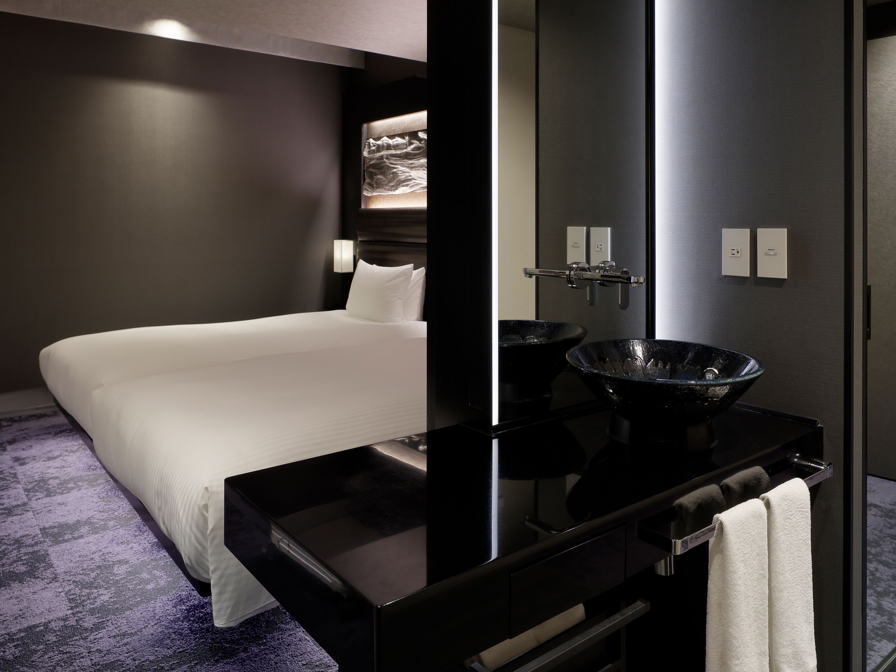 A modern hotel room with a large bed, a black vanity with a bowl sink, and a mirror.