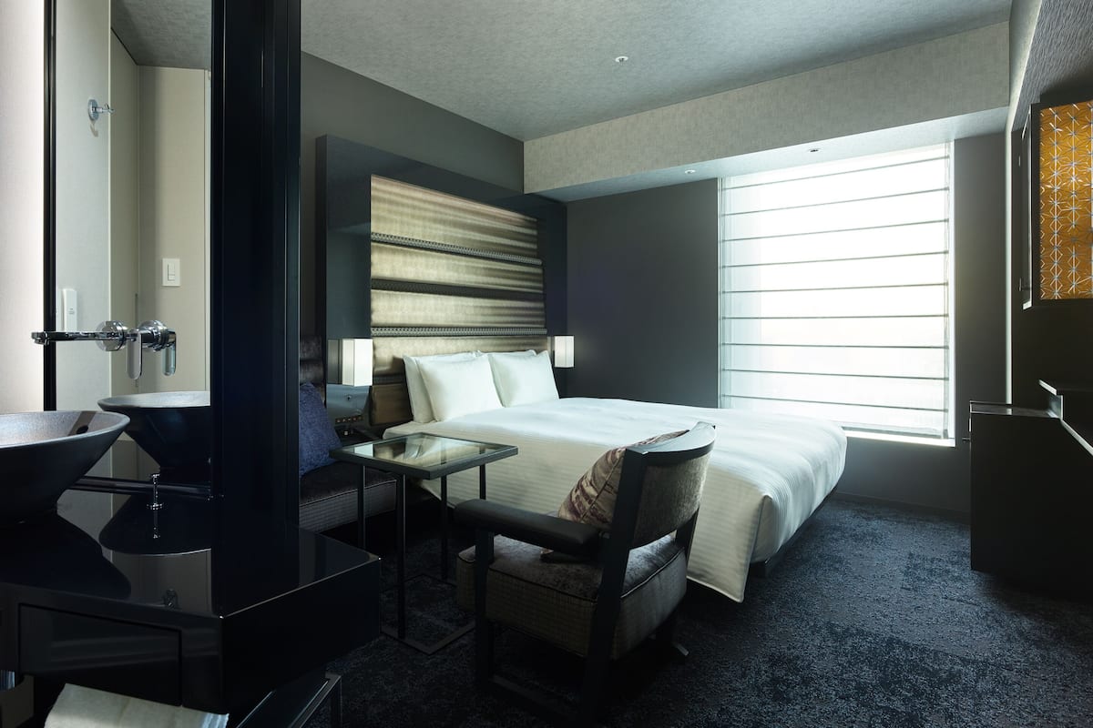 Quarto de hotel com cama, mesa de cabeceira, cadeira e banheiro com pia e espelho.