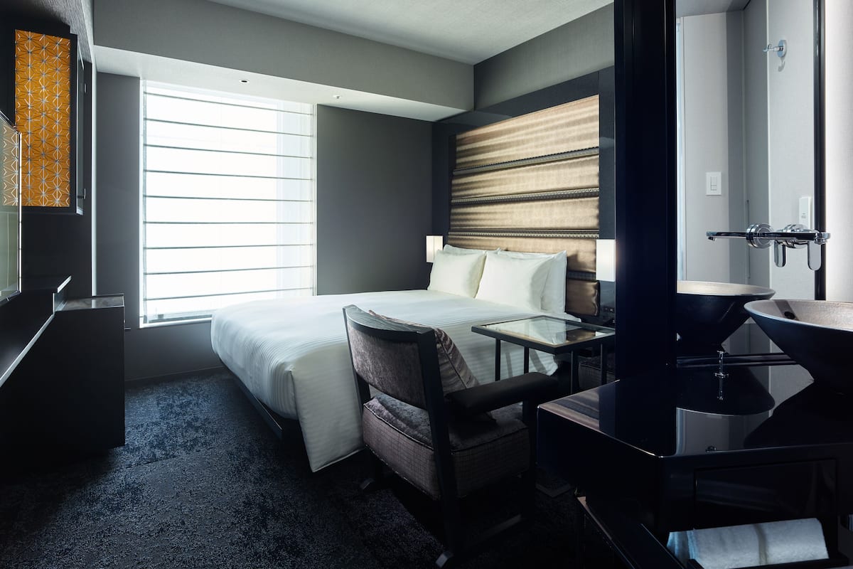 Quarto de hotel com cama, escrivaninha, cadeira, banheiro com pia e janela com persianas.