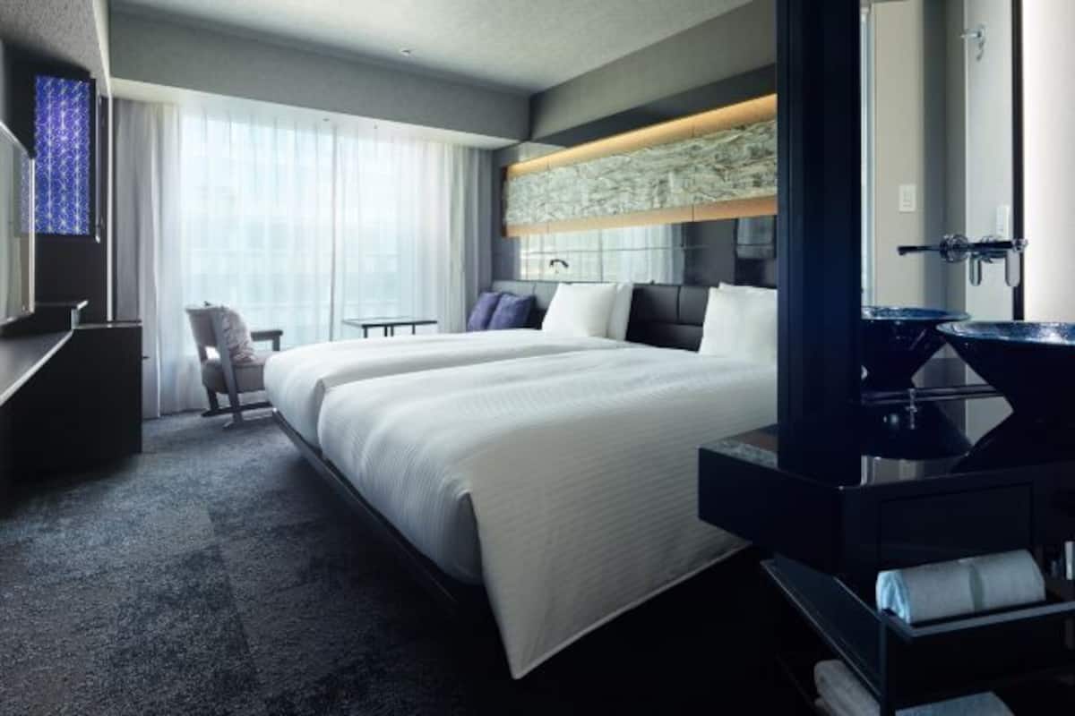 A hotel room with a large bed, a desk, a chair, a vanity with a mirror, and a window with curtains.