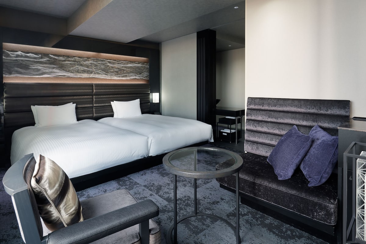 Quarto de hotel moderno com uma cama grande, uma mesa redonda, um sofá e um banquinho.