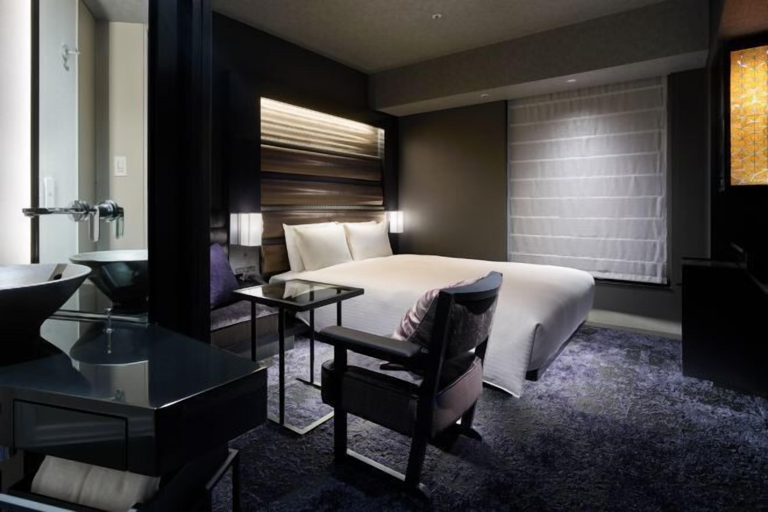 Une chambre d’hôtel équipée d’un lit, d’un bureau, d’une chaise, d’un lavabo et d’une fenêtre avec des stores.