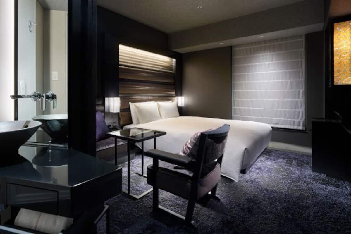 A hotel room with a bed, a desk, a chair, a sink, and a window with blinds.