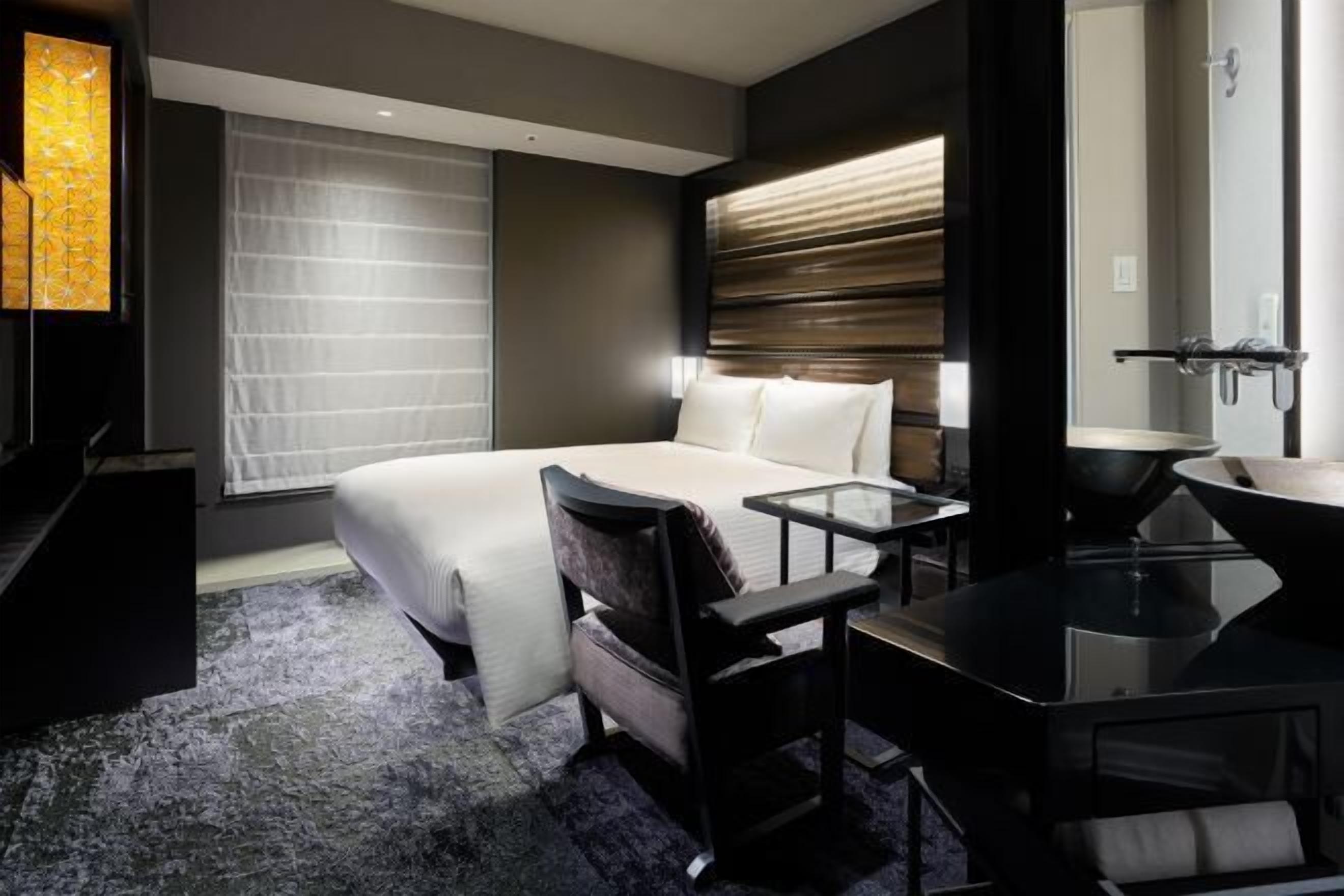 Une chambre d’hôtel avec un grand lit, un bureau avec une chaise, une salle de bain avec un lavabo et un miroir, et une fenêtre avec des stores.