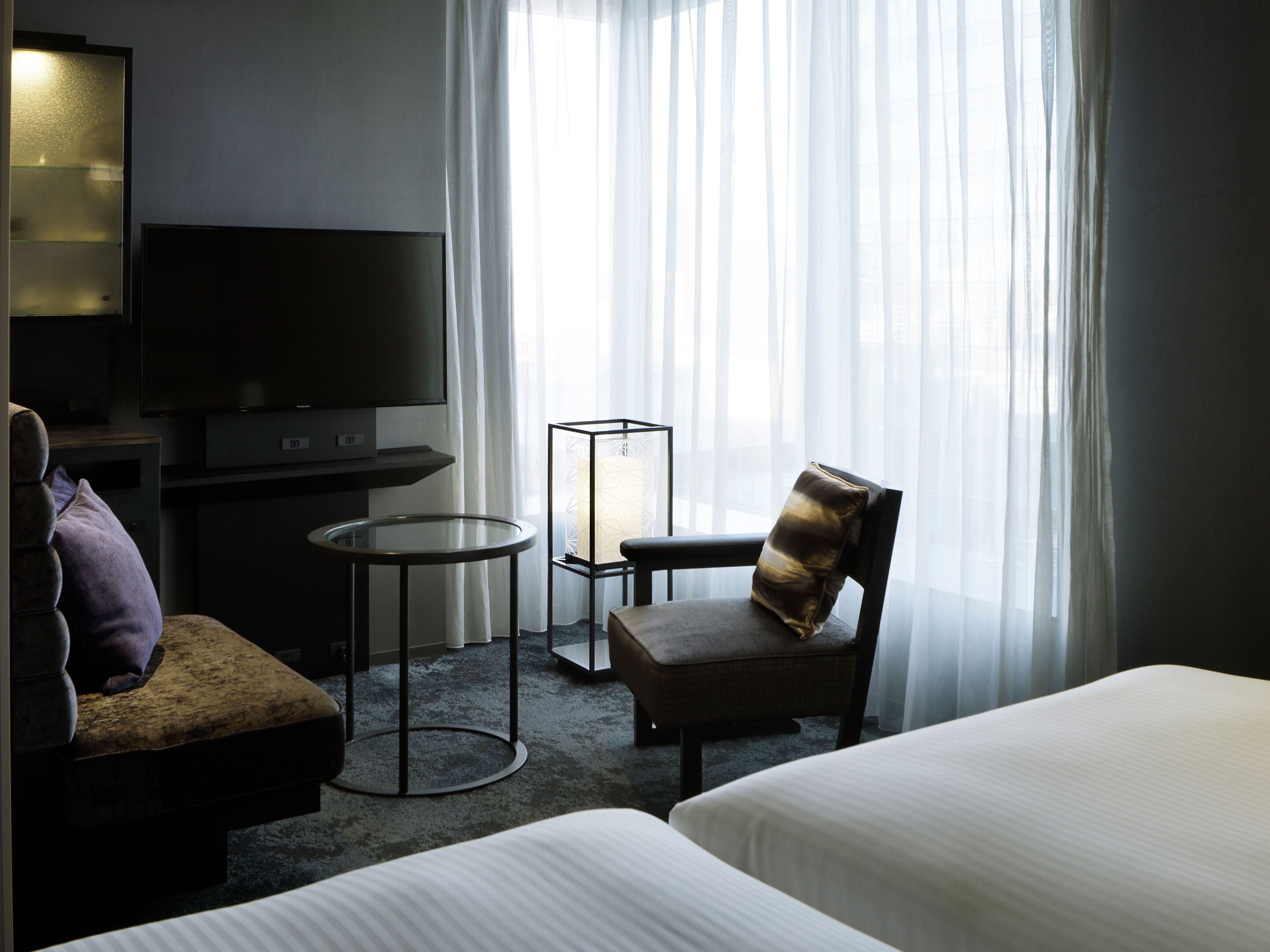 A hotel room with a bed, a chair, a small table, a TV, and a window with sheer curtains.