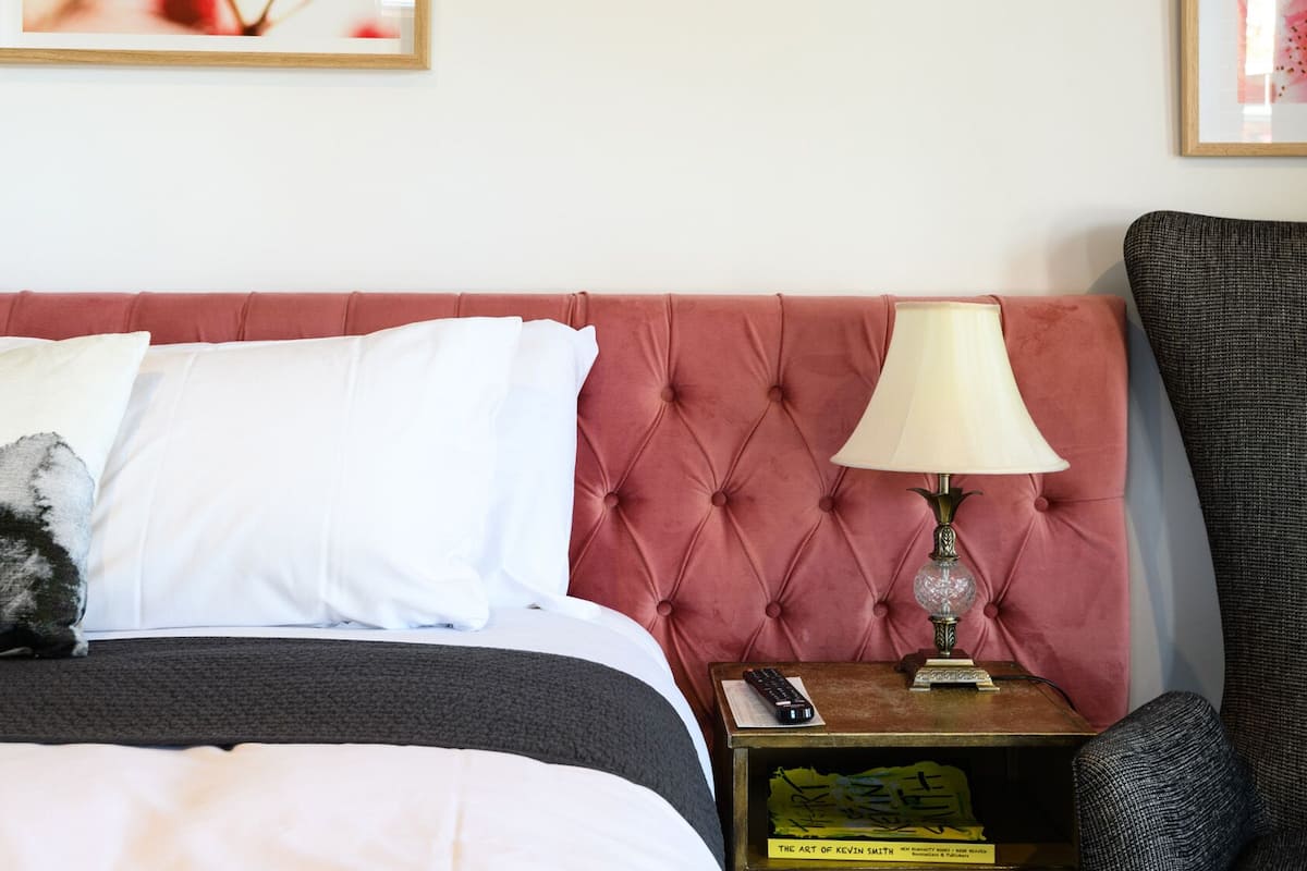 Habitación de hotel con cabecera capitonada, lámpara de noche, una silla y una mesita con un libro y un control remoto.