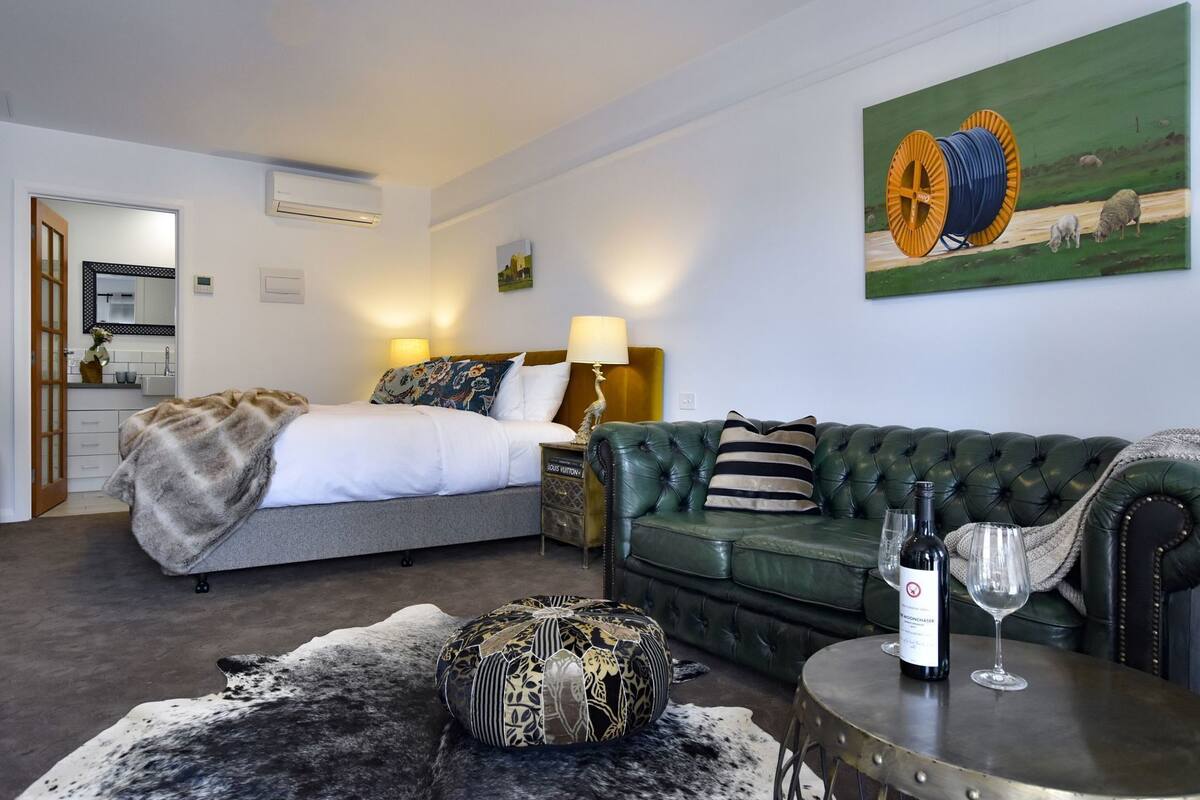 Habitación de hotel con cama, sofá verde, una mesita con una botella y un vaso, y un cuadro en la pared.